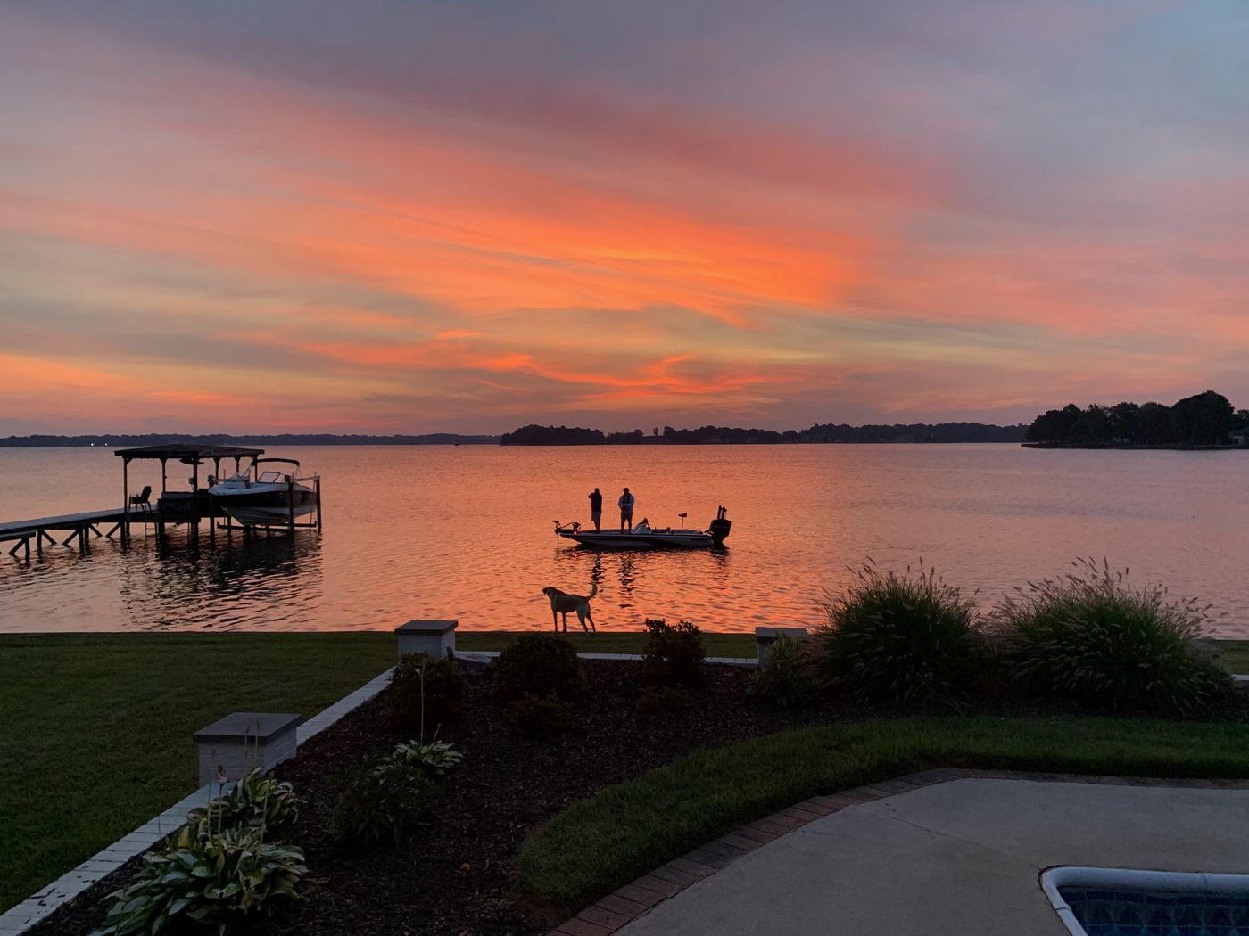 Two people fishing from a boat on Lake Norman at sunset while a dog watches from the shore